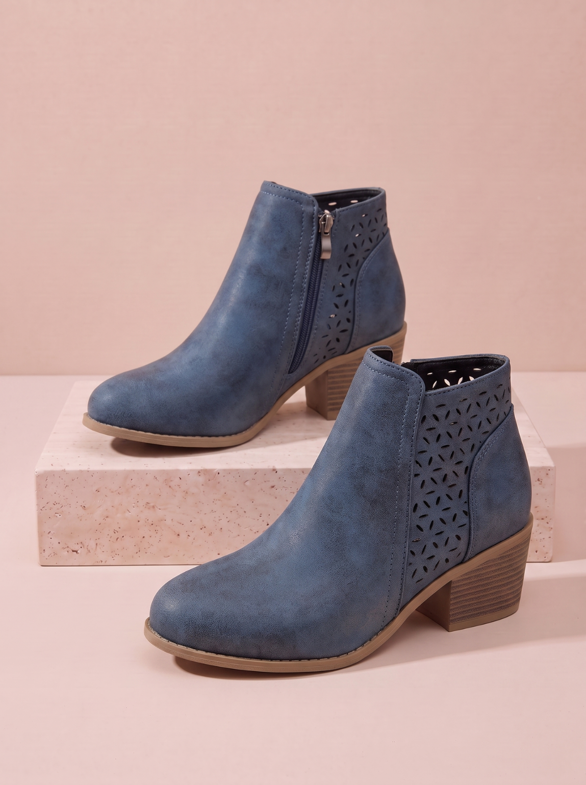 A pair of blue suede ankle boots with side cutout detailing and stacked block heels, displayed on a light pink background with a stone riser.